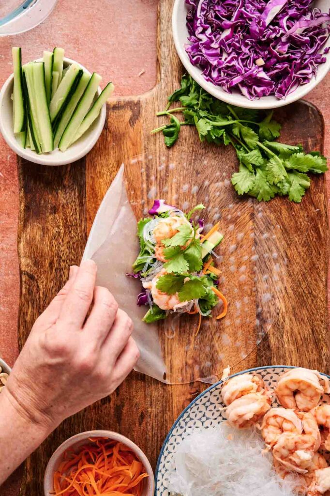 A hand rolls a Spring Roll with Shrimp in a rice paper wrapper, filled with cilantro, cucumber, shredded carrots, and purple cabbage on a wooden board, surrounded by fresh ingredients.