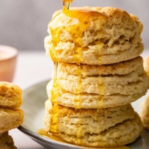 A stack of flaky sourdough biscuits on a plate is being drizzled with golden honey from above.