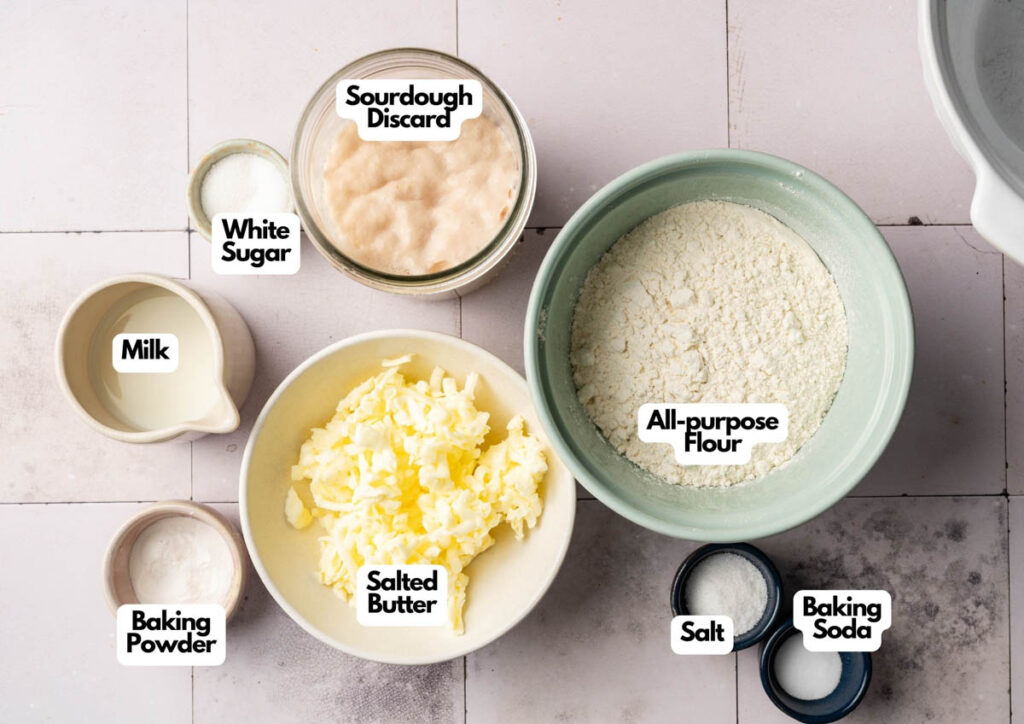 Top-down view of measured baking ingredients in bowls for Sourdough Biscuits: sourdough discard, white sugar, milk, baking powder, salted butter, all-purpose flour, salt, and baking soda on a tiled surface.