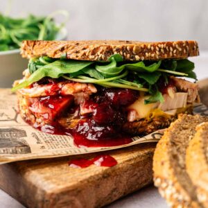 A Thanksgiving Turkey Sandwich with sliced turkey, cheese, cranberry sauce, and fresh greens on multigrain bread sits on a wooden board next to two slices of bread.