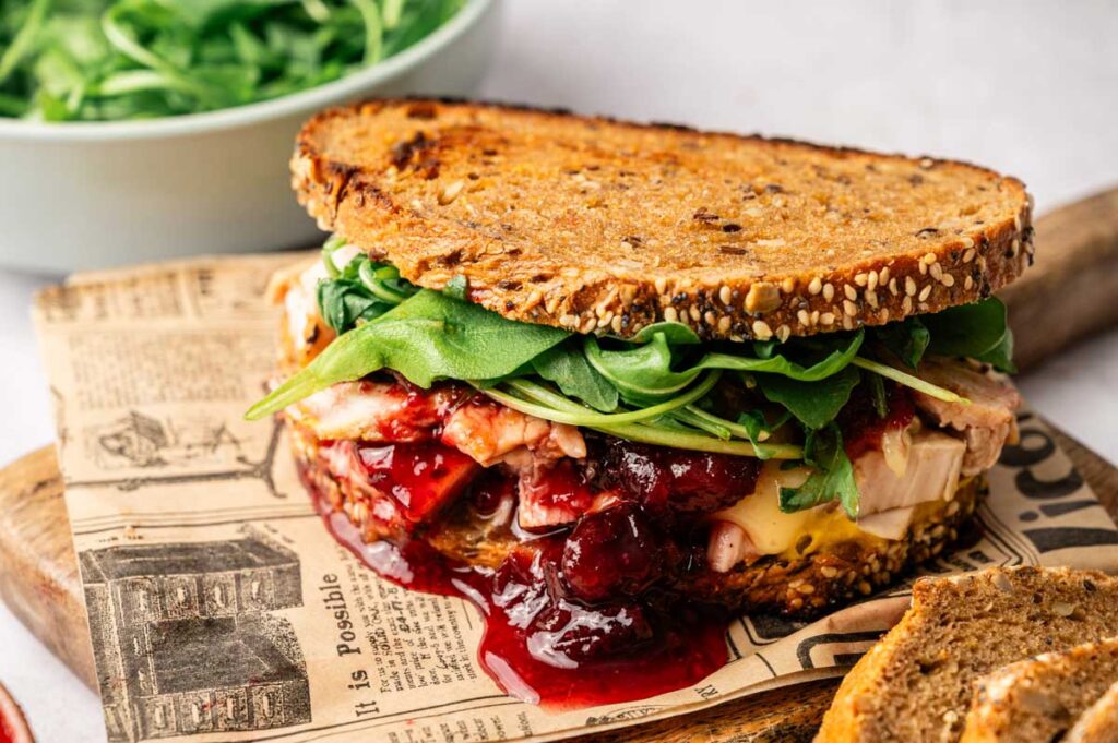 A Thanksgiving Turkey Sandwich with whole grain bread, turkey, cheese, arugula, and cranberry sauce sits on newspaper print parchment, with a bowl of arugula in the background.
