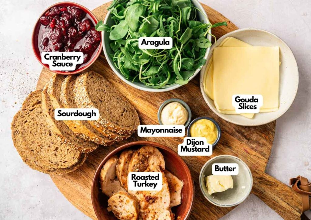 A wooden board with labeled ingredients for a Thanksgiving Turkey Sandwich: cranberry sauce, arugula, gouda slices, sourdough, mayonnaise, Dijon mustard, butter, and roasted turkey.