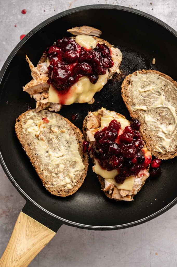 Three slices of whole grain bread in a skillet, two topped with Thanksgiving Turkey Sandwich favorites&mdash;turkey, melted cheese, and cranberry sauce&mdash;and one slice spread with butter.