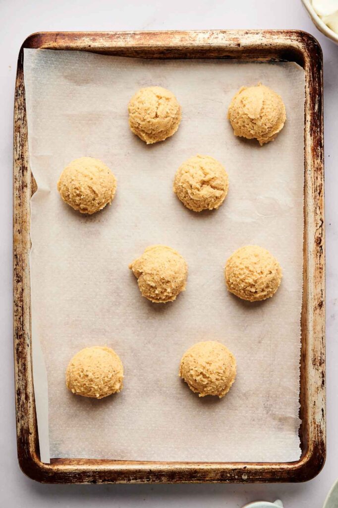 A baking sheet lined with parchment paper holds eight evenly spaced scoops of raw tiramisu cookie dough, ready to be baked.