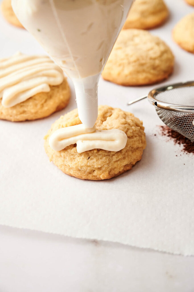 A piping bag applies white icing to a round Tiramisu Cookie on parchment paper, with other cookies and a metal sifter nearby.