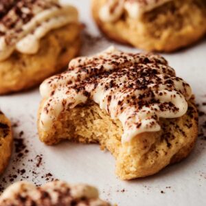 Close-up of Tiramisu Cookies topped with frosting and cocoa powder, with one cookie showing a bite taken out of it, all arranged on a white surface.