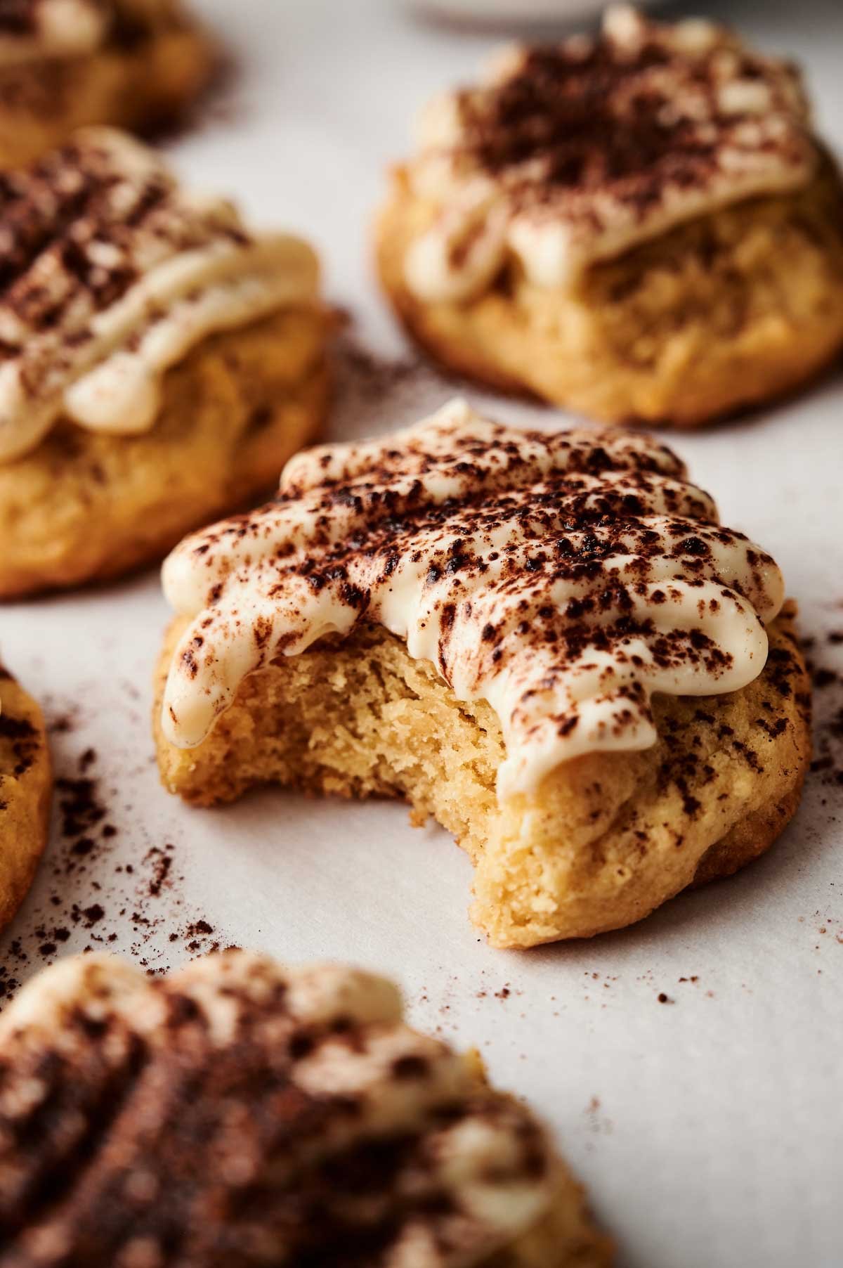 Close-up of Tiramisu Cookies topped with frosting and cocoa powder, with one cookie showing a bite taken out of it, all arranged on a white surface.