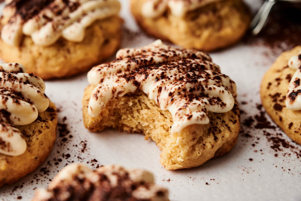 A close-up of Tiramisu Cookies topped with piped cream and sprinkled cocoa powder, with one cookie showing a bite taken out of it.