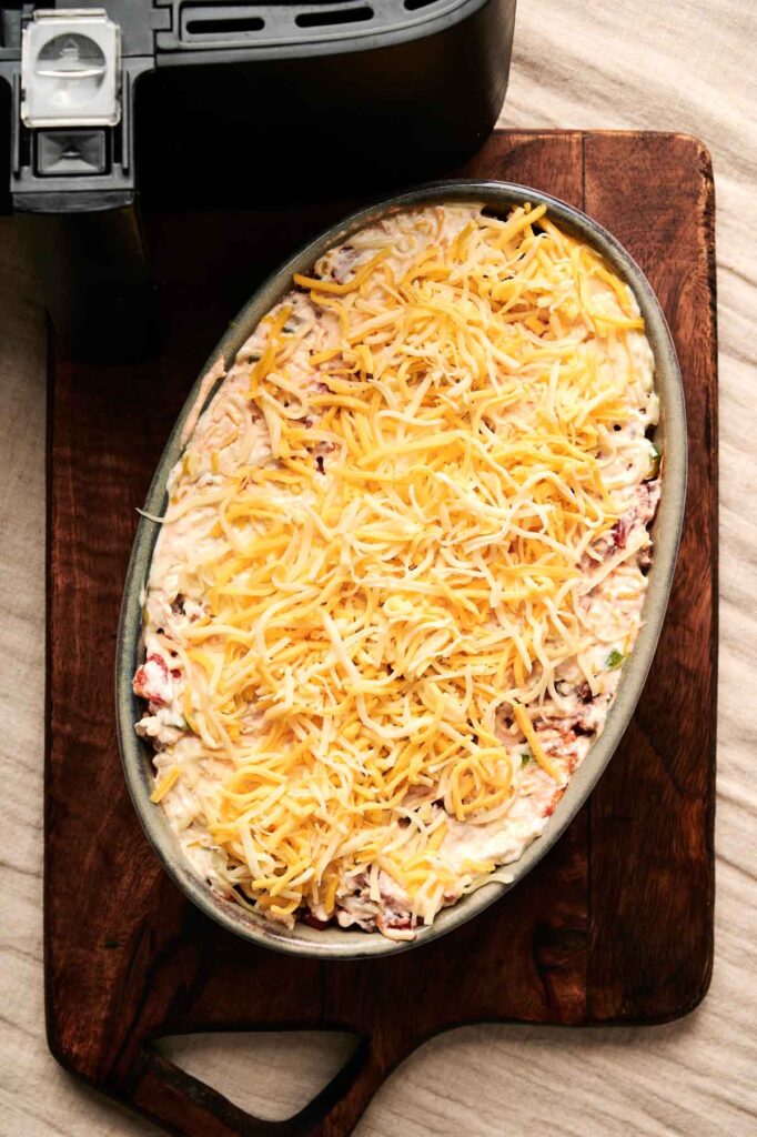 An oval baking dish filled with uncooked Air Fryer Cattle Drive Casserole sits on a wooden board next to an air fryer, ready to be cooked to cheesy perfection.