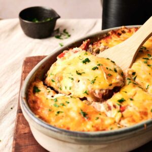 A baked Air Fryer Cattle Drive Casserole topped with melted cheese and herbs in a ceramic dish, with a wooden spatula lifting a portion.