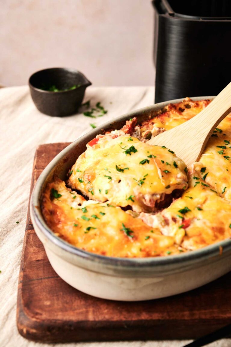 A baked Air Fryer Cattle Drive Casserole topped with melted cheese and herbs in a ceramic dish, with a wooden spatula lifting a portion.
