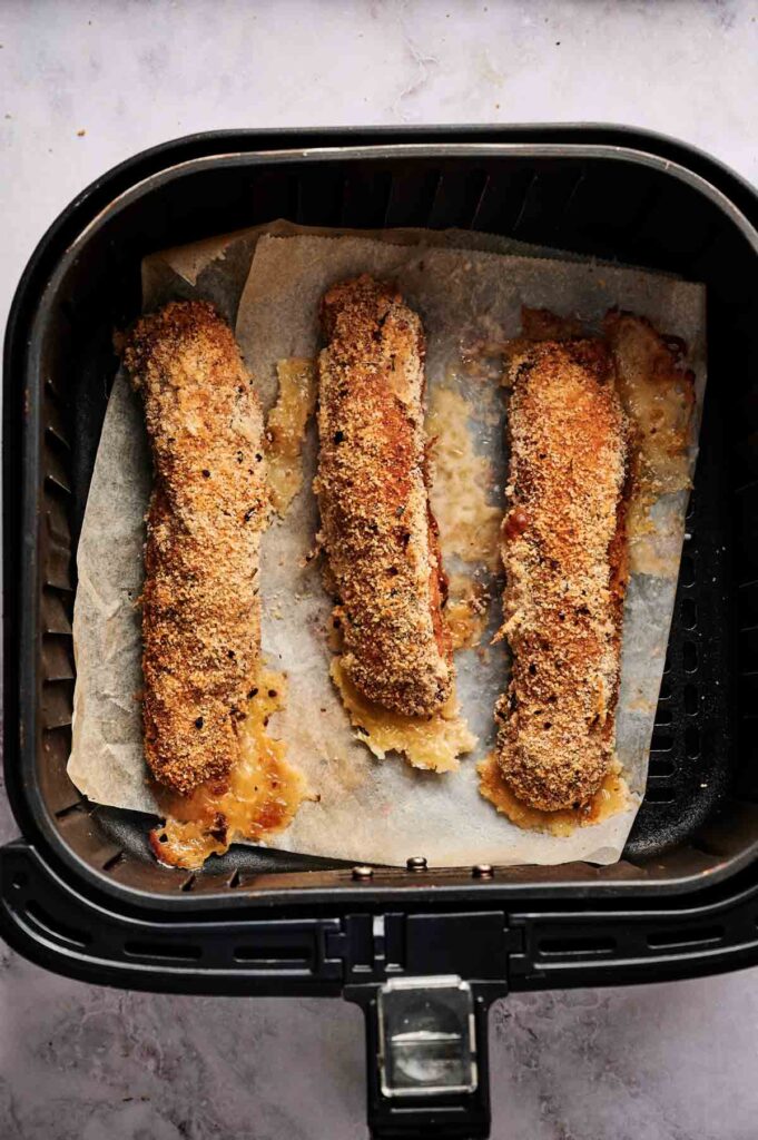 Three breaded mozzarella sticks on parchment paper inside an air fryer basket, perfect as a cheesy snack or a side to your Air Fryer Chicken Cordon Bleu.