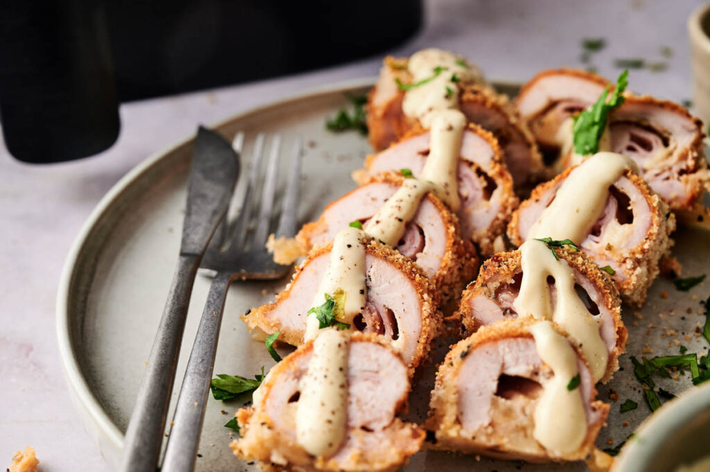 Sliced Air Fryer Chicken Cordon Bleu rolls topped with creamy sauce and herbs are arranged on a plate, with a fork and knife beside them.