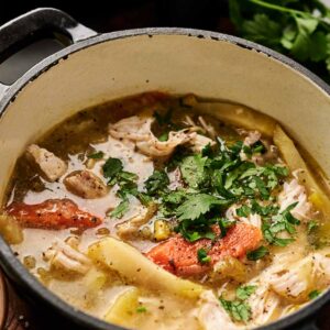 A pot of Air Fryer Chicken Soup with noodles, tender chicken chunks, tomatoes, and fresh cilantro on top, sitting on a wooden surface.