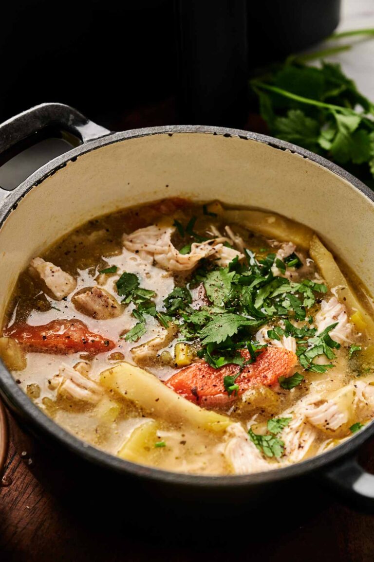 A pot of Air Fryer Chicken Soup with noodles, tender chicken chunks, tomatoes, and fresh cilantro on top, sitting on a wooden surface.
