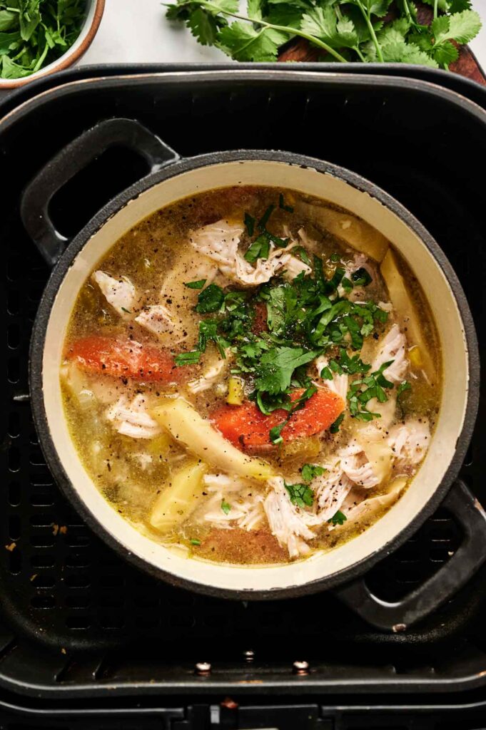 A pot of hearty Air Fryer Chicken Soup with shredded chicken, tomatoes, potatoes, and herbs sits in a black cooking appliance.