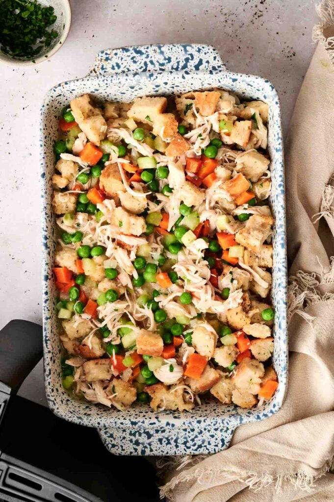 A baking dish filled with a mixture of bread cubes, shredded chicken, peas, and diced carrots, this Air Fryer Chicken and Stuffing Casserole sits on a light surface next to a beige cloth.