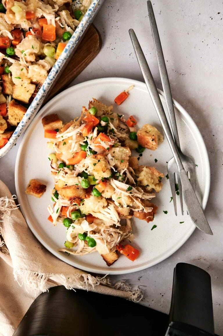 A plate of Air Fryer Chicken and Stuffing Casserole featuring bread cubes, peas, and diced carrots, served with a fork and knife on a white surface.