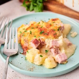 A serving of cheesy Air Fryer Ham and Potato Casserole with cubes of ham on a light blue plate, garnished with chopped parsley, with a fork and knife beside it.
