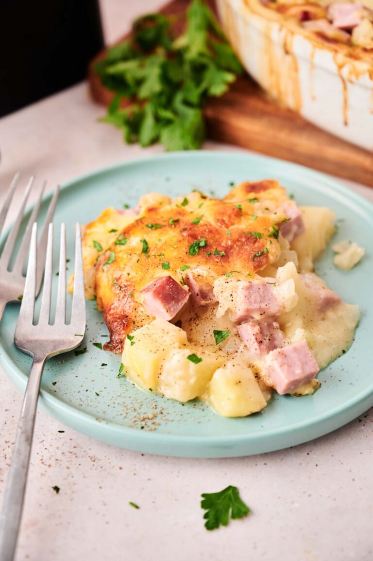A serving of cheesy Air Fryer Ham and Potato Casserole with cubes of ham on a light blue plate, garnished with chopped parsley, with a fork and knife beside it.
