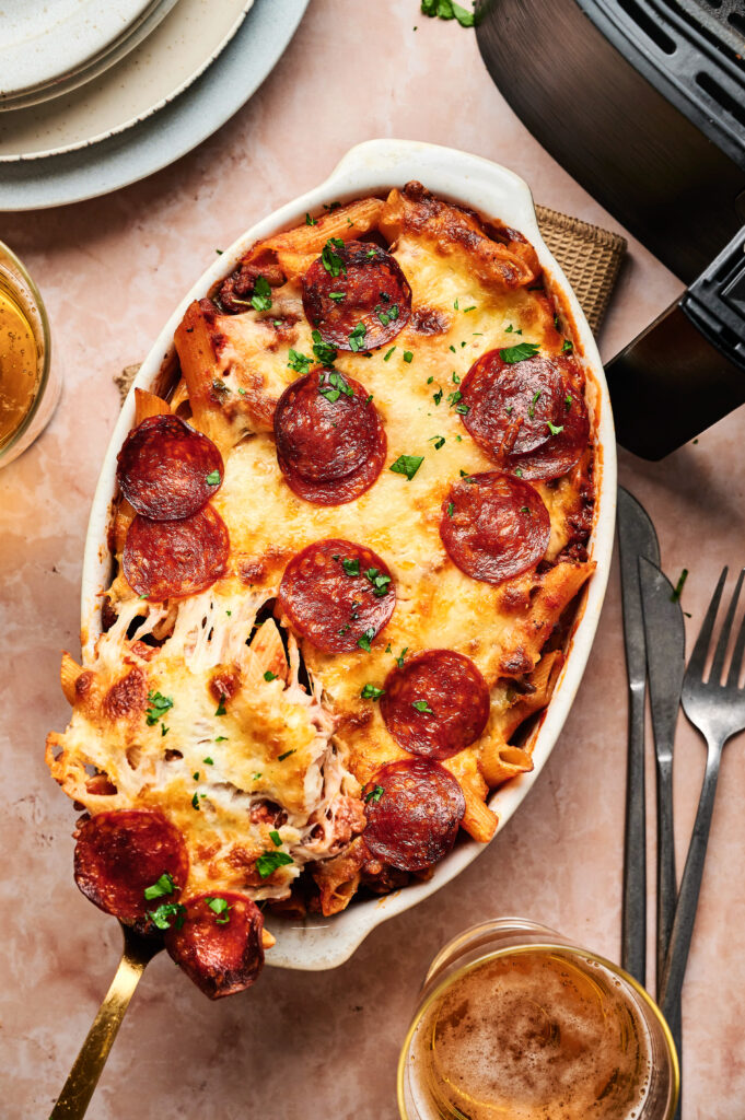 Air Fryer Pizza Casserole features baked pasta topped with melted cheese and pepperoni slices, garnished with chopped herbs, and served in an oval dish next to drinks and utensils.