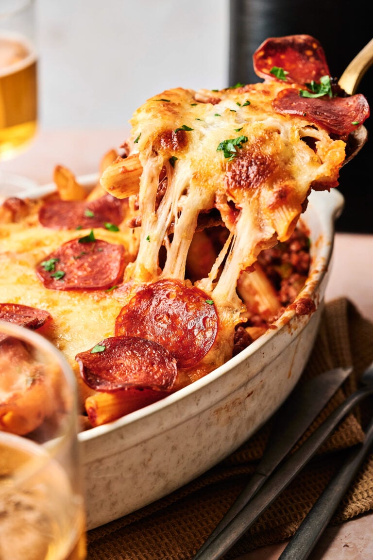An Air Fryer Pizza Casserole, topped with melted cheese and pepperoni, is being served from a white casserole dish with a serving spoon.
