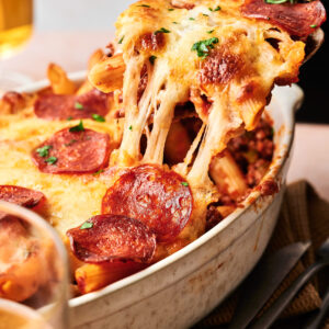 An Air Fryer Pizza Casserole, topped with melted cheese and pepperoni, is being served from a white casserole dish with a serving spoon.