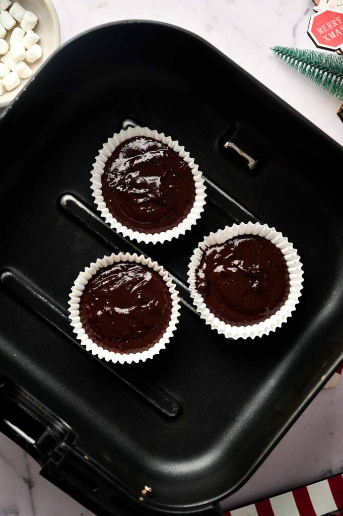 Three unbaked Air Fryer Santa Cupcakes batter portions in white paper liners are placed inside an air fryer basket on a countertop.