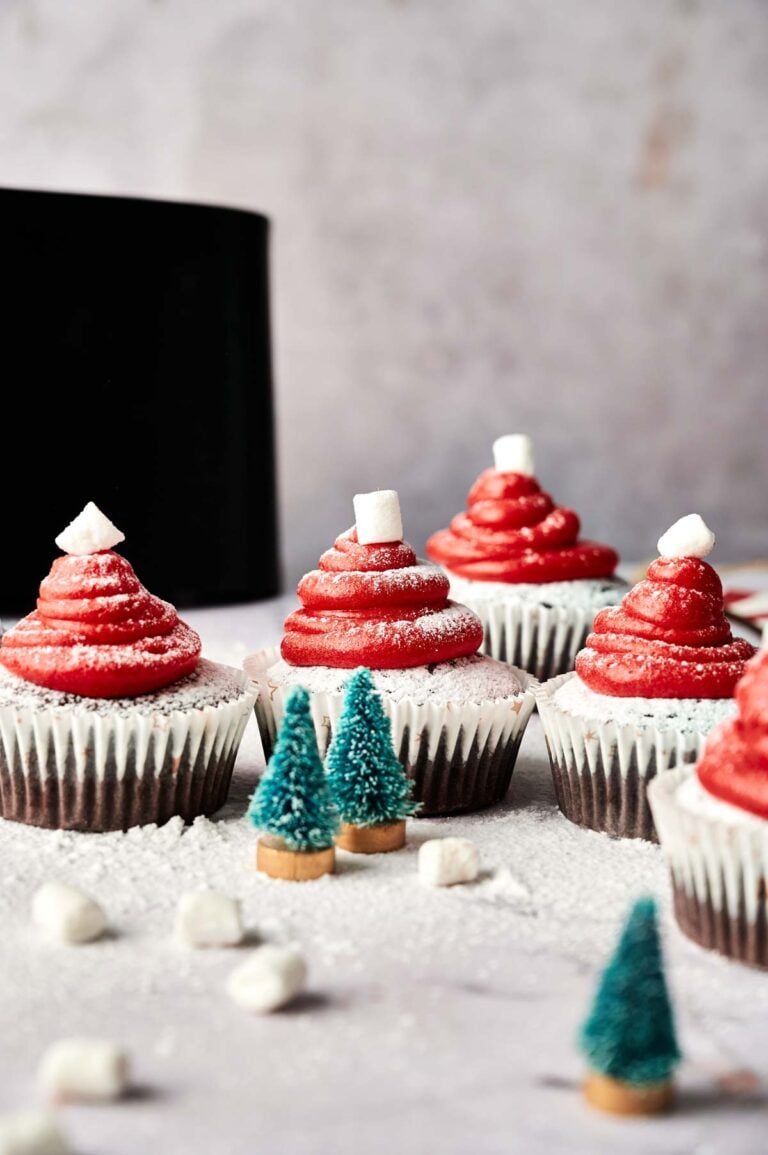 Air Fryer Santa Cupcakes with red frosting shaped like Santa hats and topped with mini marshmallows are arranged on a light surface, surrounded by small decorative trees and extra marshmallows.