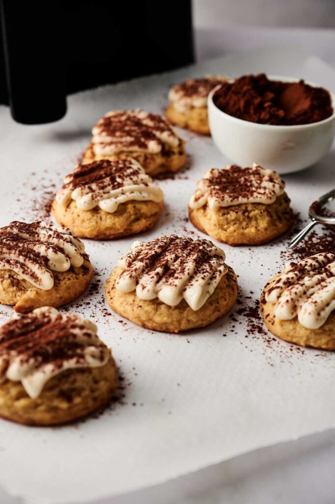 Eight Air Fryer Tiramisu Cookies topped with cream and cocoa powder are arranged on parchment paper next to a bowl of cocoa powder and a metal sifter.