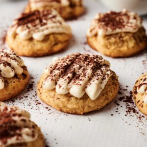 Several Air Fryer Tiramisu Cookies topped with piped cream and dusted with cocoa powder are arranged on a sheet of parchment paper. A small bowl of cocoa powder is in the background.