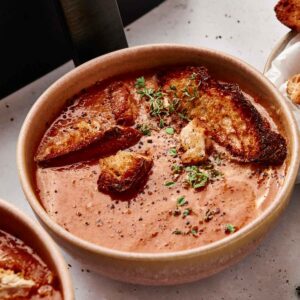 A bowl of Air Fryer Tomato Soup topped with croutons, cracked pepper, and fresh herbs sits on a light-colored surface.