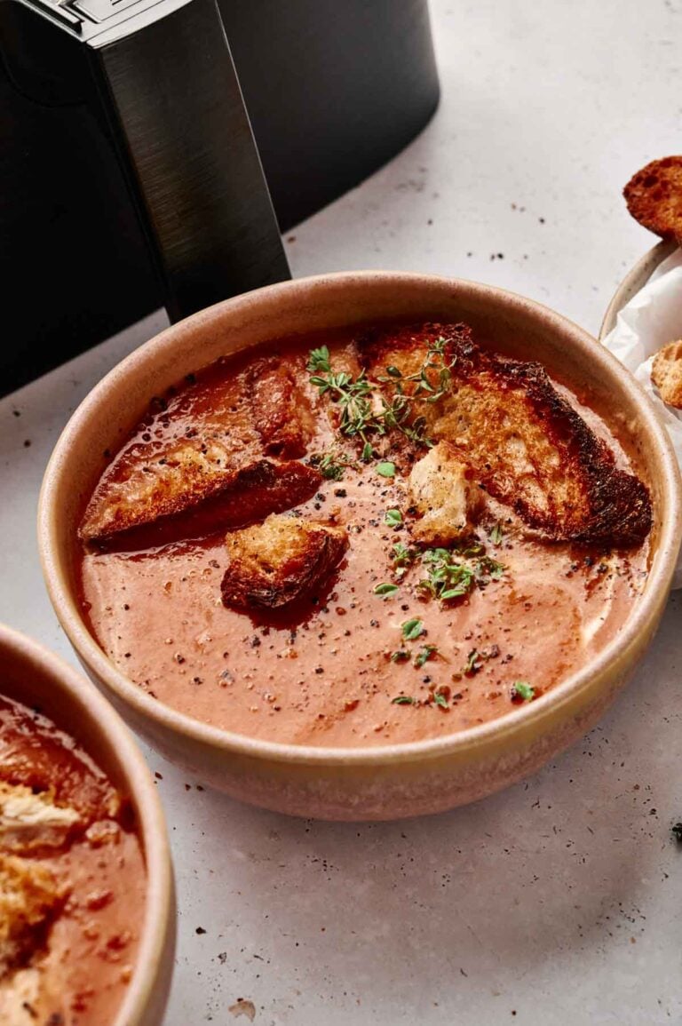 A bowl of Air Fryer Tomato Soup topped with croutons, cracked pepper, and fresh herbs sits on a light-colored surface.
