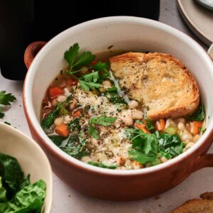 A bowl of Air Fryer White Bean Soup topped with fresh parsley and a slice of toasted bread, served alongside greens and extra bread for a wholesome, comforting meal.