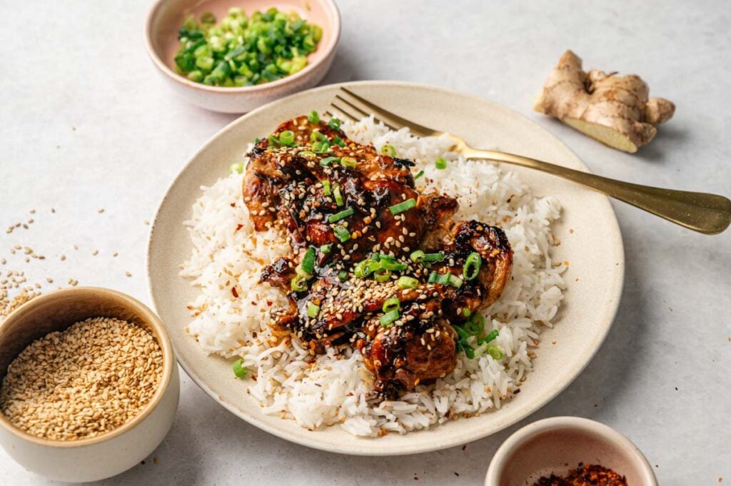 A plate of Asian Chicken Thighs glazed to perfection, topped with sesame seeds and chopped green onions, served over white rice, with bowls of sesame seeds, green onions, and ginger nearby.