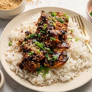A plate of white rice topped with glazed Asian Chicken Thighs, garnished with sesame seeds and chopped green onions. A fork rests on the side of the plate.