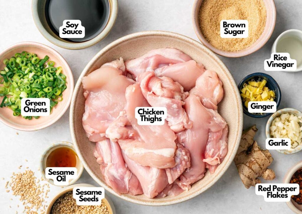A bowl of raw Asian Chicken Thighs is surrounded by labeled bowls of soy sauce, brown sugar, rice vinegar, green onions, ginger, garlic, red pepper flakes, sesame oil, and sesame seeds.