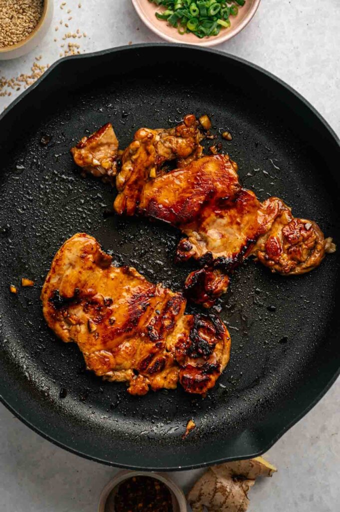 Two cooked pieces of Asian Chicken Thighs are shown in a black skillet, with some sauce residue and oil visible around them.