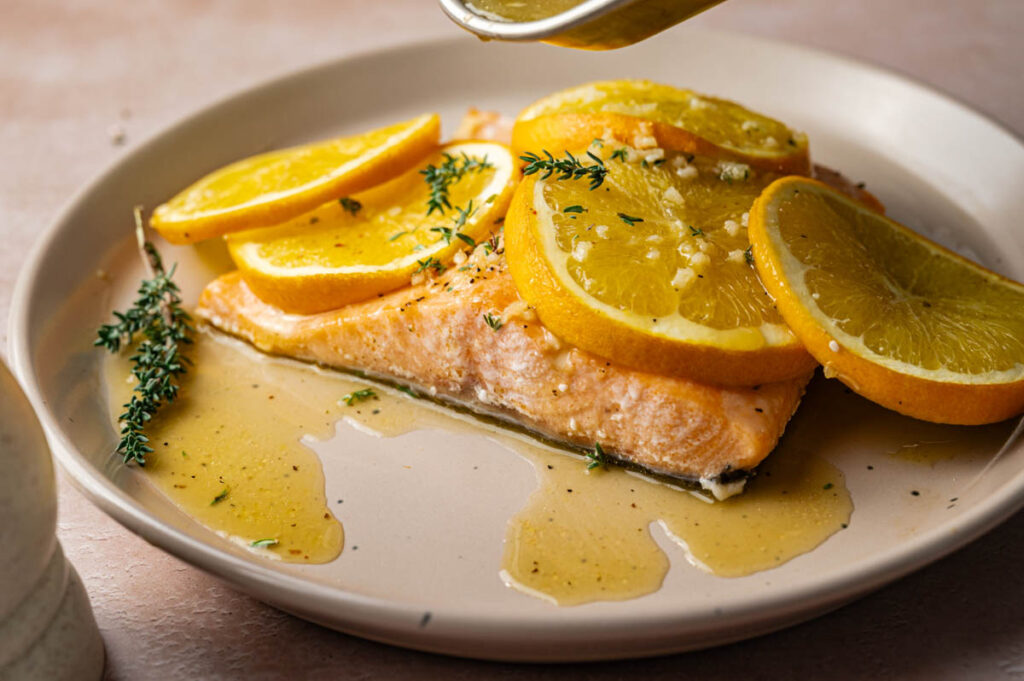 A baked salmon fillet topped with orange slices and herbs is drizzled with a light sauce on a beige plate.