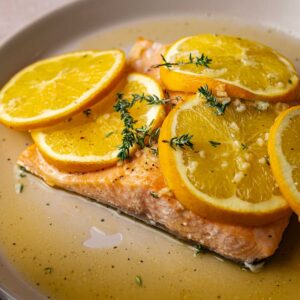 A baked salmon fillet topped with orange slices and garnished with fresh thyme sits on a beige plate with a light sauce.