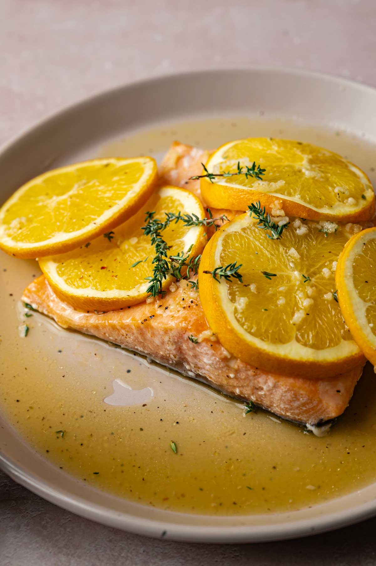 A baked salmon fillet topped with orange slices and garnished with fresh thyme sits on a beige plate with a light sauce.