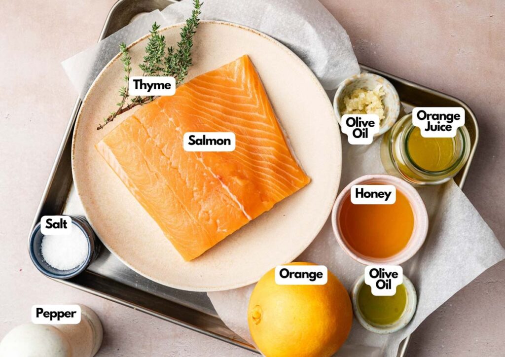 A plate with raw salmon and thyme, surrounded by bowls of salt, pepper, olive oil, orange juice, honey, and a whole orange on a tray. Each ingredient is labeled for a delicious baked salmon preparation.