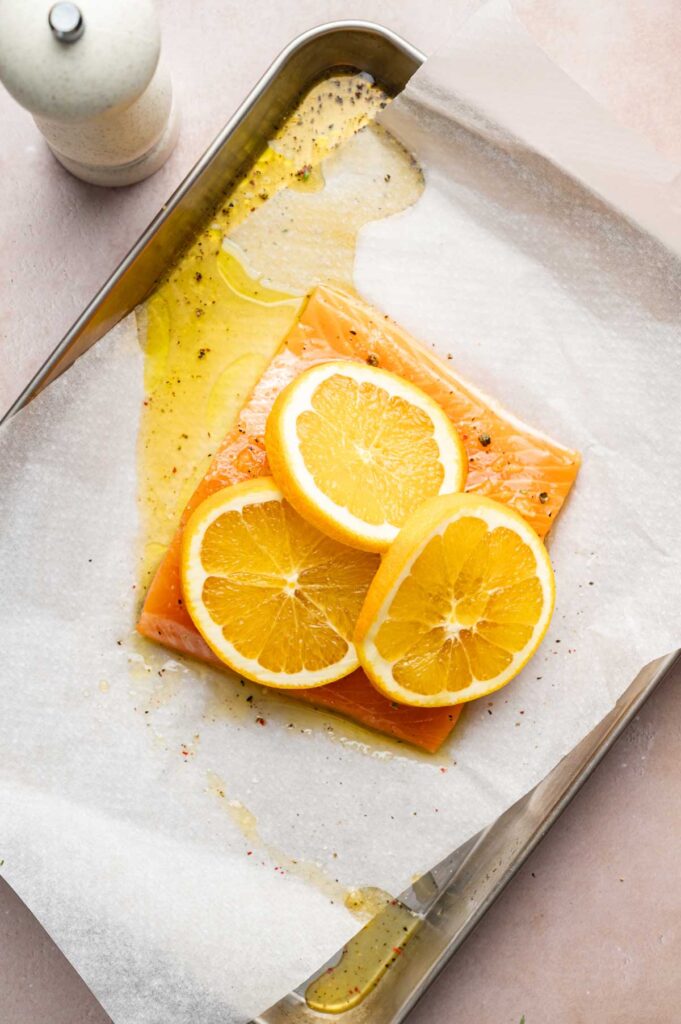 A raw salmon fillet topped with three lemon slices sits on parchment paper in a baking tray, ready to become delicious baked salmon, drizzled with oil and sprinkled with black pepper.