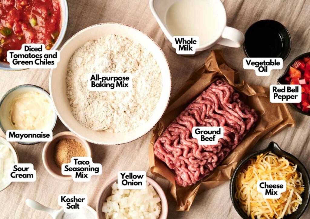 Ingredients for Air Fryer Cattle Drive Casserole arranged on a table, including ground beef, cheese mix, baking mix, diced tomatoes with green chiles, red bell pepper, onion, milk, oil, mayonnaise, sour cream, and seasonings.