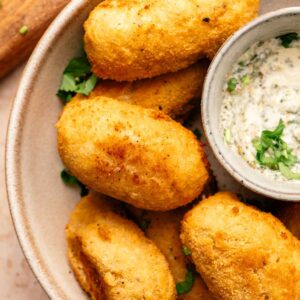 A plate of golden-brown Chicken Croquettes is served with a small bowl of creamy dipping sauce, garnished with chopped herbs.