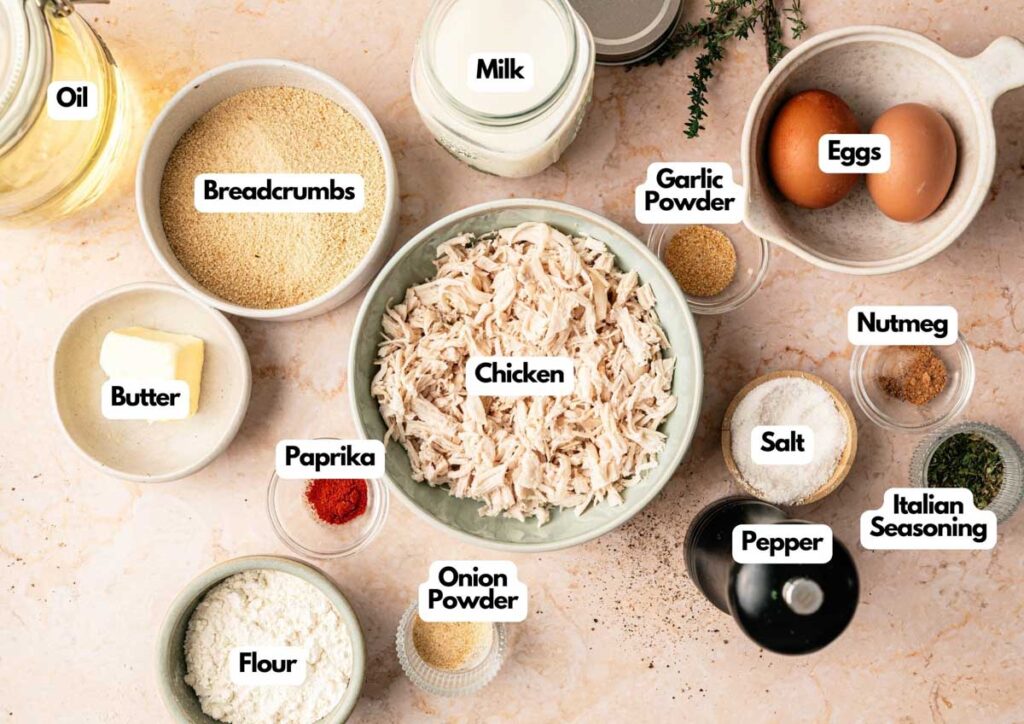 A variety of labeled ingredients for cooking Chicken Croquettes—chicken, eggs, milk, oil, butter, flour, breadcrumbs, and assorted seasonings—arranged neatly on a countertop.