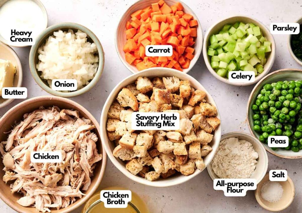 Bowls containing labeled ingredients for an Air Fryer Chicken and Stuffing Casserole, including shredded chicken, vegetables, broth, flour, butter, cream, and peas, are arranged on a light surface.