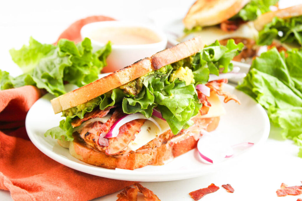 A Chipotle Chicken Club sandwich on a white plate with layers of lettuce, bacon, cheese, red onion, and tomato, served with a dipping sauce in the background.