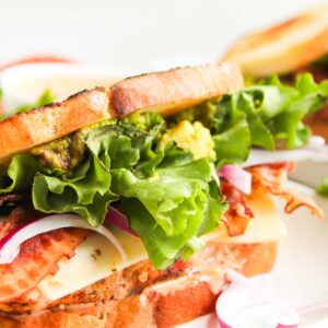 A close-up of a Chipotle Chicken Club sandwich with lettuce, bacon, cheese, red onion, and sauce between two slices of toasted bread, served on a white plate.