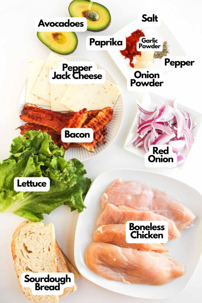 Ingredients for Chipotle Chicken Clubs displayed on a white surface, including avocado, cheese, bacon, red onion, lettuce, sourdough bread, boneless chicken, and various seasonings.